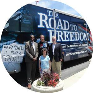 A group of four people, three gentleman and one lady, pose in front of the Road to Freedom Tour Bus during the ADA25 Celebration tour in May 2015